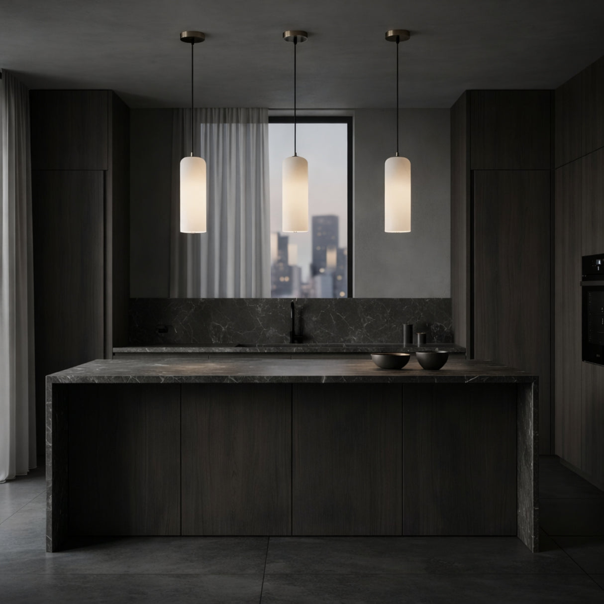 A modern kitchen features dark wood cabinets, a marble island, and three Velmora 1 Light Slim Cylindrical Alabaster Single Pendant Lights in Soft White & Brass. A dusk cityscape is visible through the window behind the sink; two bowls sit on the countertop.