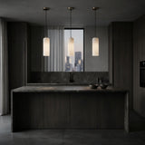 A modern kitchen features dark wood cabinets, a marble island, and three Velmora 1 Light Slim Cylindrical Alabaster Single Pendant Lights in Soft White & Brass. A dusk cityscape is visible through the window behind the sink; two bowls sit on the countertop.