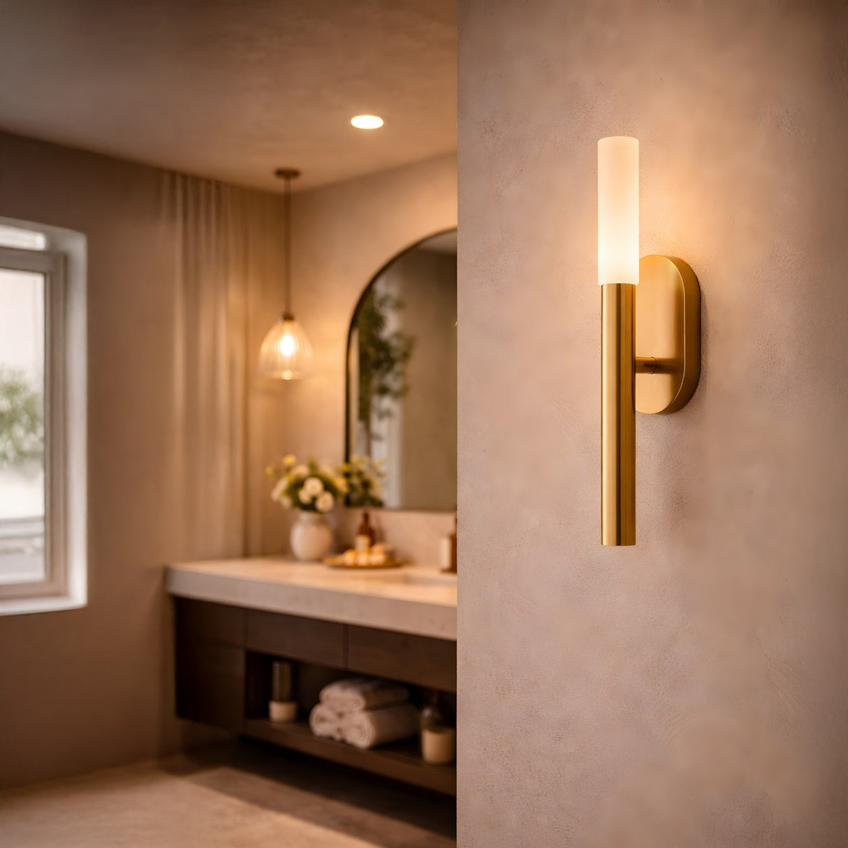 A modern bathroom features the Aurelian LED Vertical Column Alabaster Wall Light in brushed brass and soft white, a curved mirror, a pendant light, flowers on the counter, folded towels on open shelves, and natural light from a window.