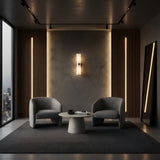A modern lounge with two gray armchairs, a round white coffee table, and Elaris 2 Light Double Cylinder Alabaster Wall Lights in soft white & brass accenting a textured wall. Large windows reveal a city view; a tall mirror leans against wood paneling.
