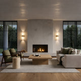 Modern living room with a central fireplace, beige walls, large windows, light wood floors, a cream sofa with a throw, two green armchairs, a wooden coffee table, and warm lighting from the Elaris 2 Light Double Cylinder Alabaster Wall Light – Soft White & Brass.