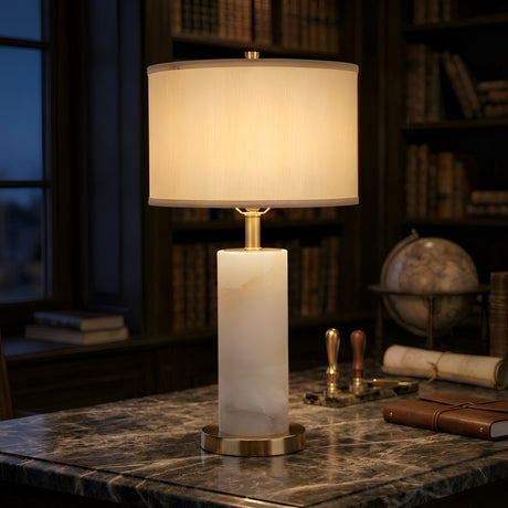 The Calynor 1 Light Column Alabaster Table Lamp - Brass & Soft White sits on a marble desk in a library, surrounded by books, a globe, and rolled-up papers.