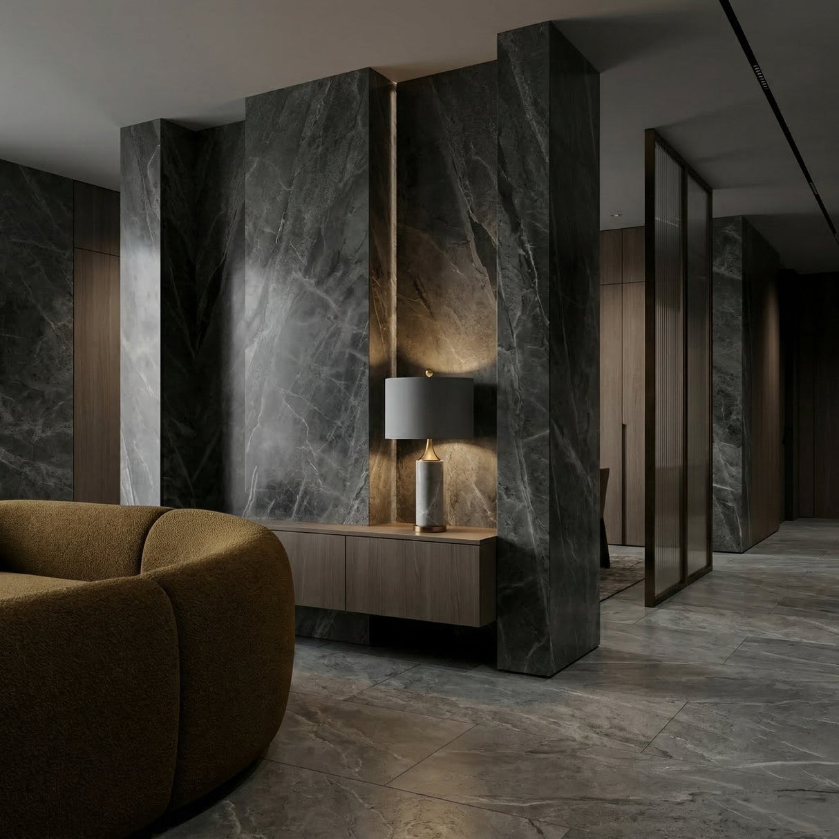 A modern interior with dark marble walls and floor, a brown curved sofa, a wooden console table, and the Valoma 1 Light Sculpted Marble Table Lamp in brass and grey, creating a sleek and luxurious atmosphere.