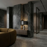 A modern interior with dark marble walls and floor, a brown curved sofa, a wooden console table, and the Valoma 1 Light Sculpted Marble Table Lamp in brass and grey, creating a sleek and luxurious atmosphere.