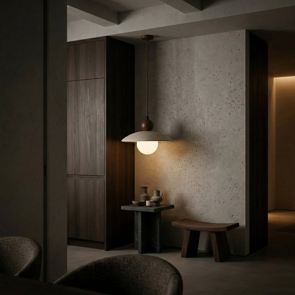A modern, minimalist interior features the Trevano 1 Light Dome Travertine Single Pendant Light in Walnut Brown & Light Beige above a small stone table with ceramic cups, a wooden stool, and textured neutral walls for a cozy atmosphere.