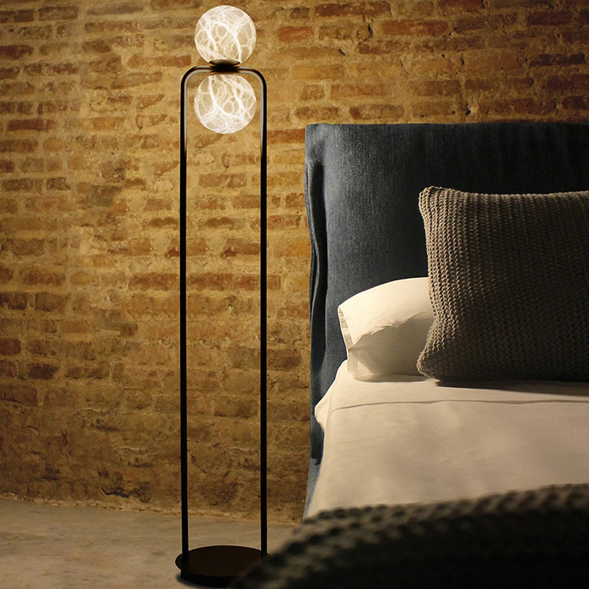 Halvessa LED 2 Light Alabaster Floor Lamp in black, tall inverted U-shaped iron frame with glowing globes, shown beside a bed with dark upholstered headboard.