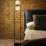 Halvessa LED 2 Light Alabaster Floor Lamp in black, tall inverted U-shaped iron frame with glowing globes, shown beside a bed with dark upholstered headboard.
