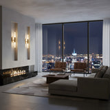 A modern living room features a beige sectional, two leather chairs, minimal decor, a fireplace, and a large window with city views. Cozy ambiance is set by the Aurelian LED Double Column Alabaster Wall Light in brushed brass & soft white.