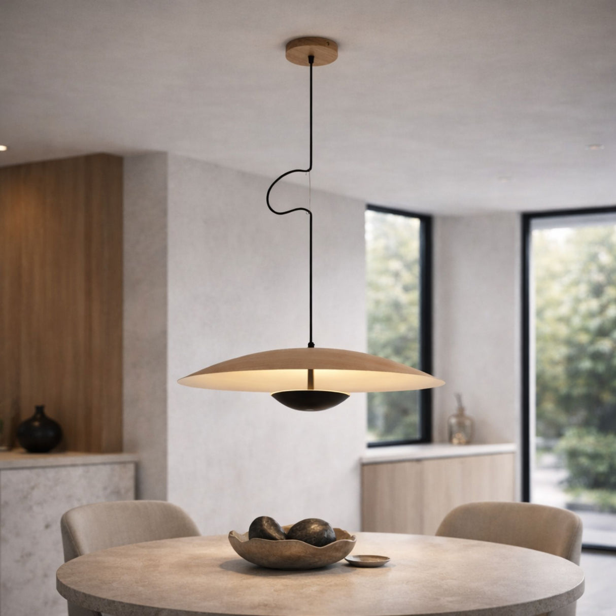 The Ginger LED Large Pendant Light - Black & Brown, with its wide flat shade, hangs above a round dining table with a decorative bowl. The neutral-toned room features large windows and minimalist decor.