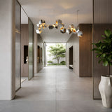 A modern, minimalist hallway with gray floors, wood-paneled walls, and the Spar Linear Suspension Light in white and gold adds elegance. Geometric pendant lighting highlights the space, while glass doors reveal a courtyard with a green tree.