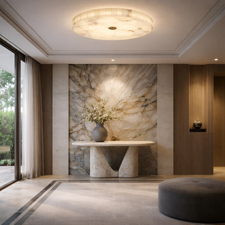 Elegant entryway with a round marble table, white flowers, and a dramatic marble wall. The Veltrix LED Large Round Alabaster Flush Ceiling Light in Soft White & Brushed Brass casts gentle light, creating a calm, sophisticated mood as sunlight enters through large windows.