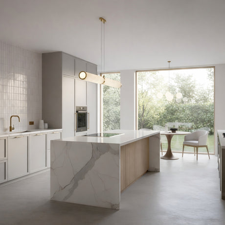 Modern kitchen with a marble island, minimalist white cabinets, built-in oven, and gold fixtures. The Oria LED Small Linear Alabaster Suspension Light in soft white & brass hangs above the round dining table near windows overlooking a green garden.