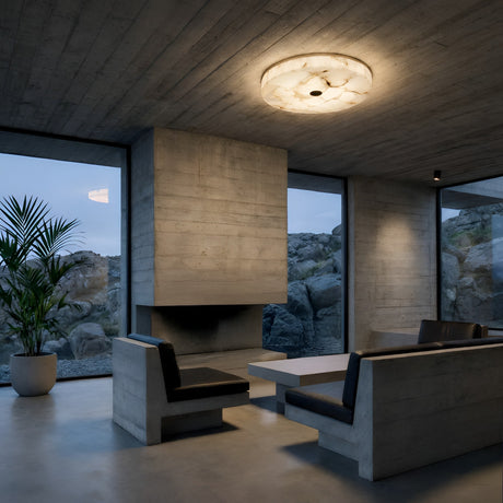 A modern, minimalist living room with concrete walls and ceiling, built-in fireplace, black and gray bench seats around a white table, large windows with rocky views, and a Veltrix LED Small Round Alabaster Flush Ceiling Light in soft white & brushed brass.