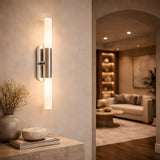 The Aurelian LED Double Column Alabaster Wall Light in Satin Nickel & Soft White glows on a textured beige wall, softly illuminating a cozy living room with a sofa, round coffee table, and built-in shelves in the background.