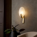 Mica 1 Light Disc Alabaster Wall Light in soft white with a curved brushed brass bracket, illuminated on a grey wall beside a plant and armchair.