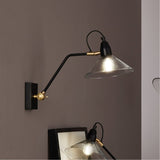 Canton Matt Black Brass & Glass Cone Wall Light with adjustable arm and clear glass shade, mounted on a taupe wall above a bedside mirror.