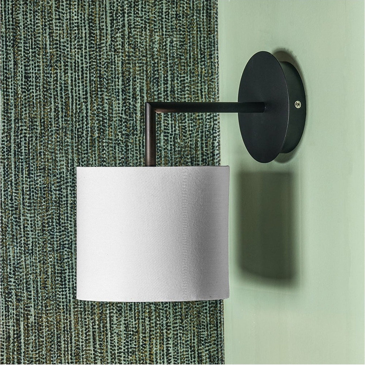 Elin Matt Black & Ivory Wall Light - 26cm, with a round matt black backplate and arm holding an ivory fabric drum shade, mounted on a green textured wall.