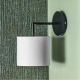Elin Matt Black & Ivory Wall Light - 26cm, with a round matt black backplate and arm holding an ivory fabric drum shade, mounted on a green textured wall.