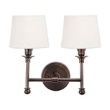 Piero Antique Bronze Double Wall Light with two tapered white fabric shades on curved metal arms, mounted on a round bronze backplate, shown on a plain white background.