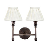 Piero Antique Bronze Double Wall Light with two pleated cream fabric shades on curved arms, mounted on a round backplate, shown on a plain white background.