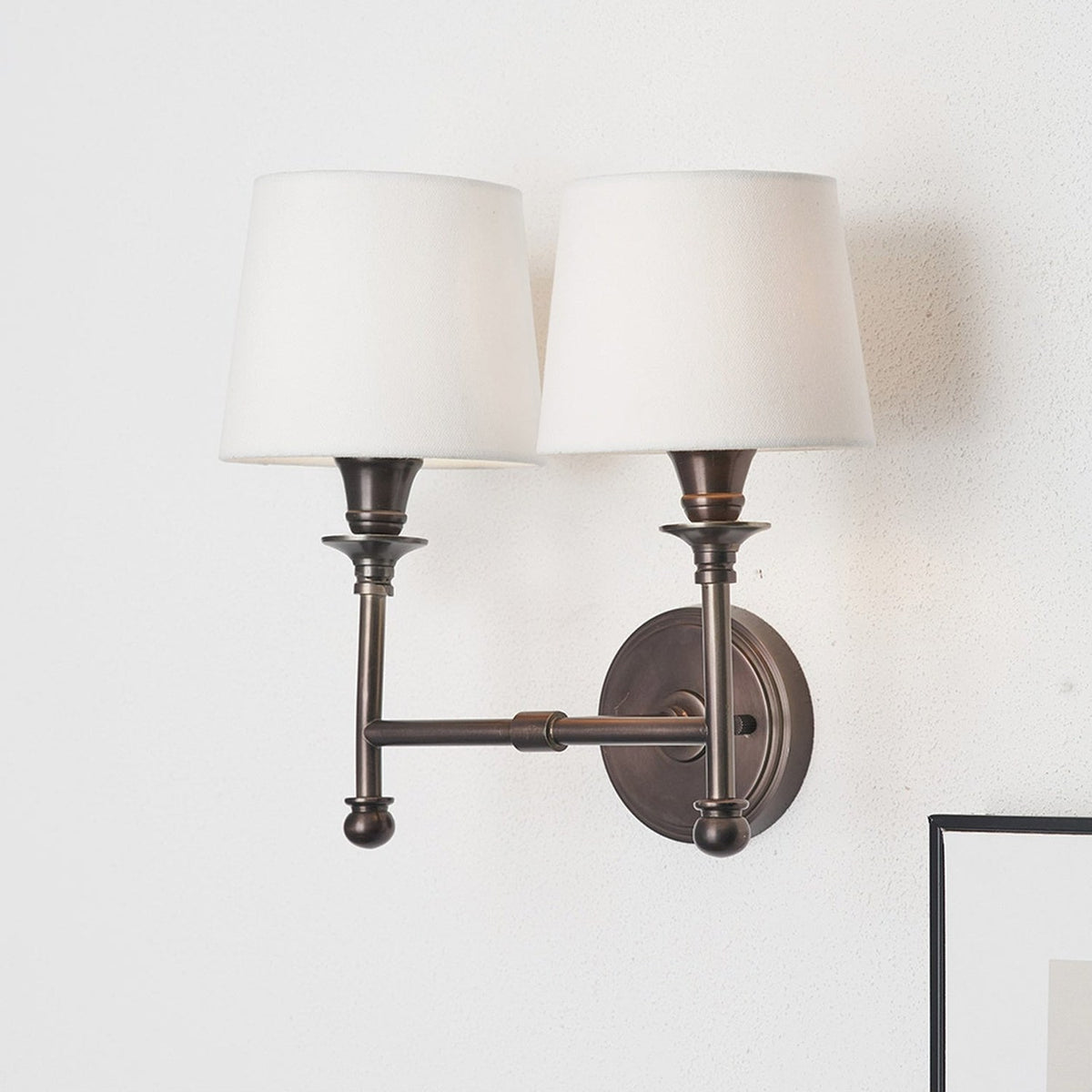 Piero Antique Bronze Double Wall Light with two arms and cream fabric shades, mounted on a plain white wall.
