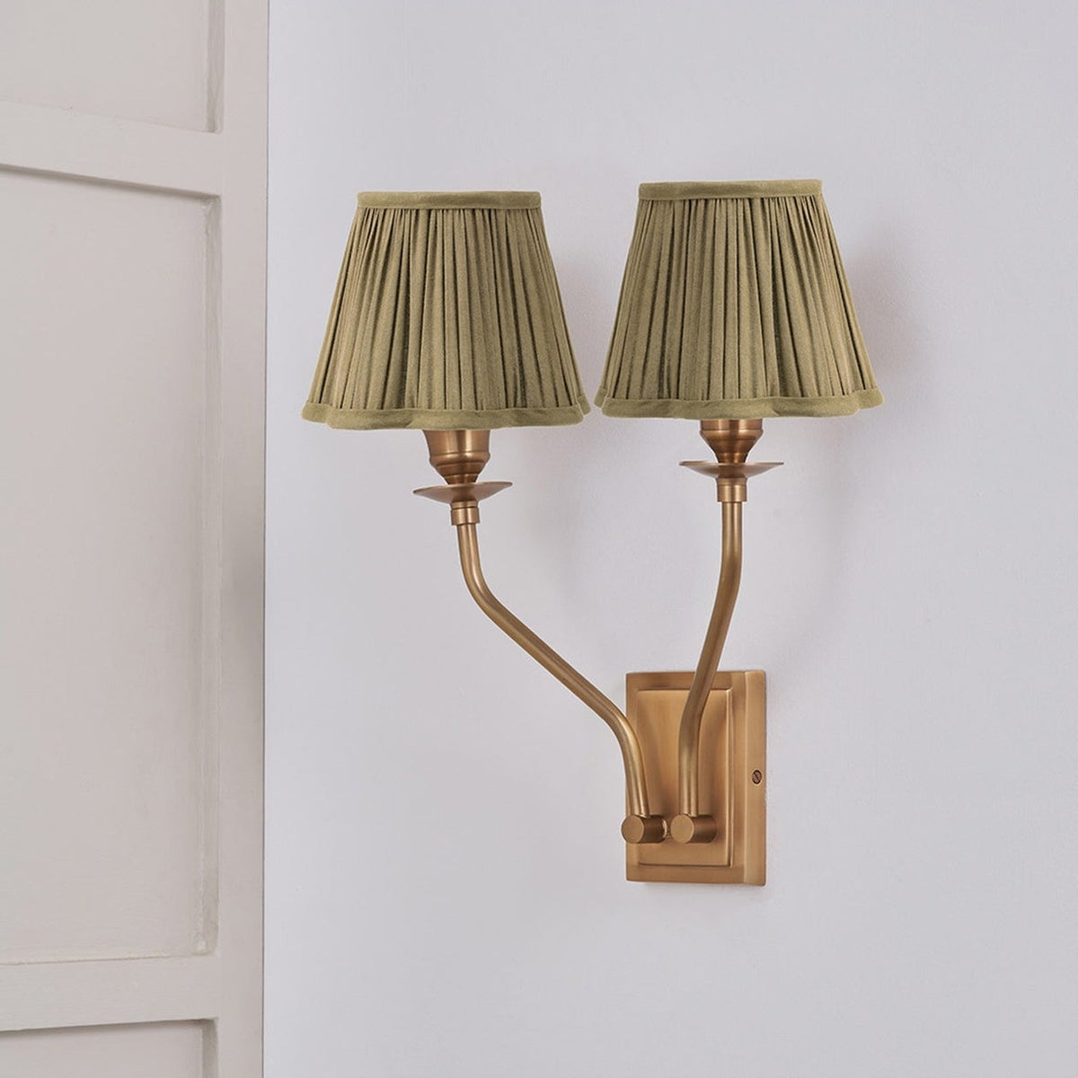 Tatiana antique brass double wall light with two curved arms and pleated olive green fabric shades, mounted on a light grey wall beside a white panelled door.