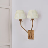 Tatiana Antique Brass Double Wall Light with two cream silk mix pleated shades, mounted on a light grey wall beside a white panelled door.