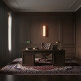 A classic wooden office features a large desk with a green banker’s lamp, books, and a laptop. An executive chair sits behind the desk, while an Almora LED Medium Cylinder Alabaster Wall Light adds sophistication above the decorative rug.