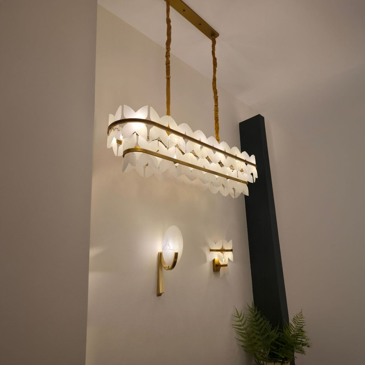 Petraia Floral Alabaster Wall Light in brushed brass with layered stone petals, shown lit on a wall beside a matching leaf-form sconce and pendant.