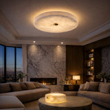 A modern living room with marble walls, beige sofas, a fireplace, bookshelves, large windows showing a cityscape at dusk, and an Aleron LED Extra Small Round Alabaster Flush Ceiling Light in Soft White & Brushed Brass.