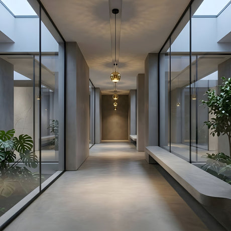 A modern hallway with large glass walls, minimalistic design, indoor plants, concrete benches along the sides, and natural light from skylights. The space is illuminated by Grand Ball Glass Pendant 25cm - Gold lights suspended overhead.