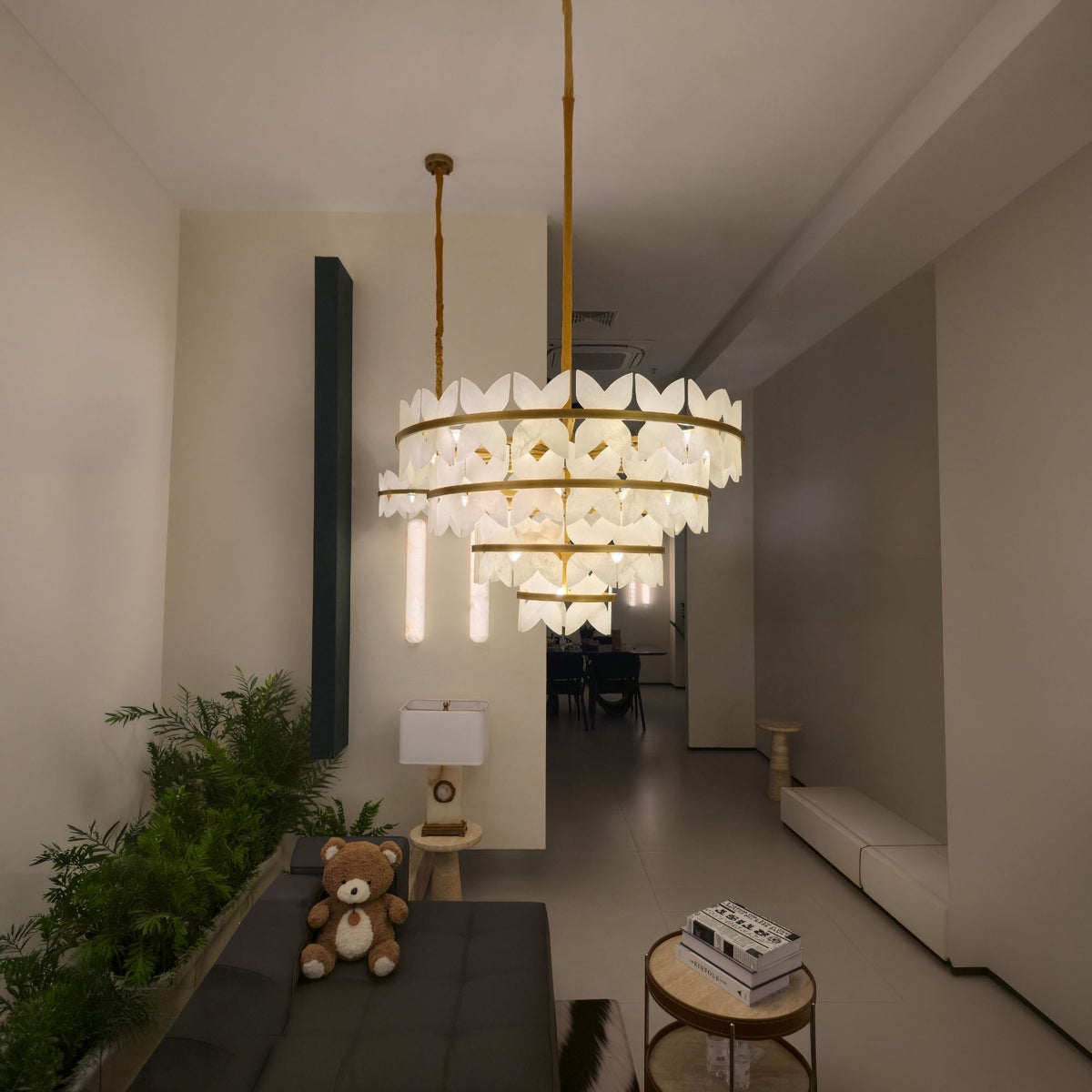 Petraia 4-tier chandelier with sculpted alabaster petals on brushed brass rings, illuminated in a hallway setting near a living area with plants and books.
