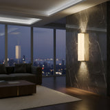 A modern living room with a city view at dusk, a dark marble accent wall featuring the Valmere LED Small Linear Alabaster Wall Light in soft white, a brown sofa, and a wooden coffee table on a light striped rug.