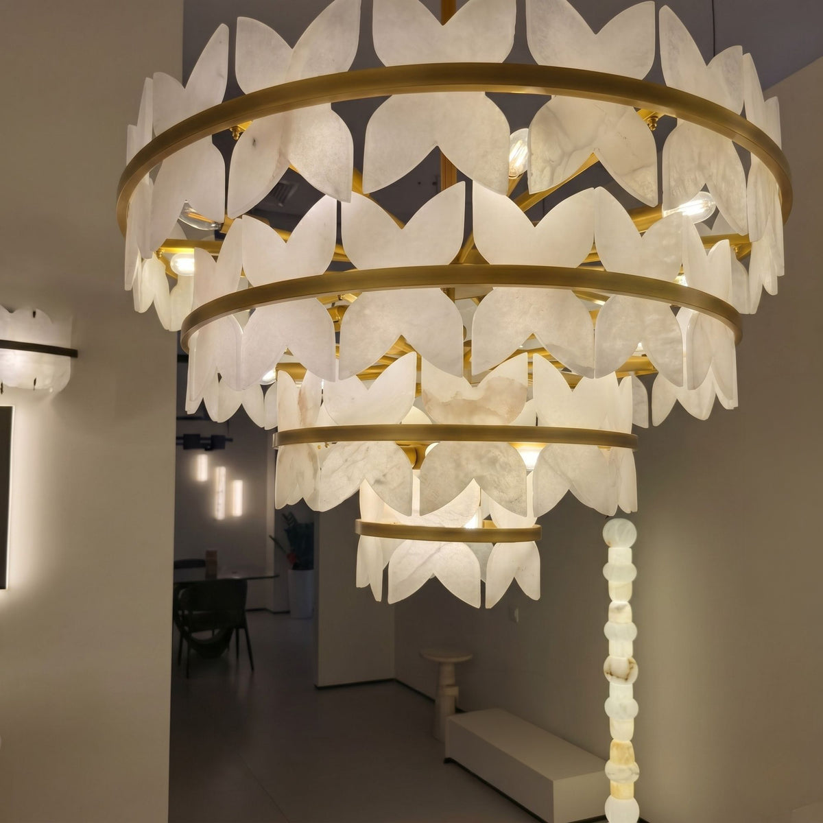 Petraia 4-tier floral alabaster chandelier with brushed brass rings, carved petal-like leaves and warm integrated lighting, shown installed in a hallway setting.
