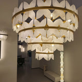 Petraia 4-tier floral alabaster chandelier with brushed brass rings, carved petal-like leaves and warm integrated lighting, shown installed in a hallway setting.