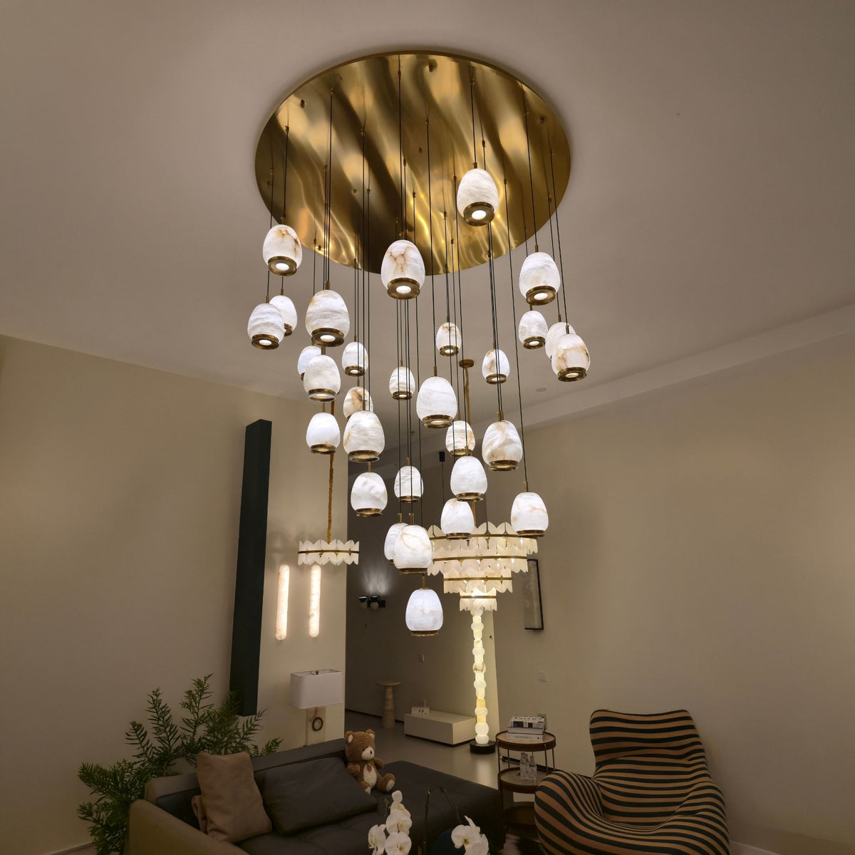 Aurevia 31-Light LED Alabaster Chandelier with brushed brass round canopy and cascading pebble-shaped shades, shown suspended in a living room setting.