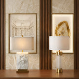 The Lunivar 1 Light Faceted Column Alabaster Table Lamp in Brass & Soft White sits on a wooden surface against a textured wall with framed art, its warm, neutral tones adding an elegant, modern touch to the space.