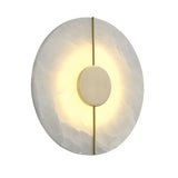 The Elaris LED Round Alabaster Wall Light in Soft White & Brass features a circular alabaster base, central brass disc, and vertical brass rod, softly glowing from behind to cast a radiant halo on the stone surface.
