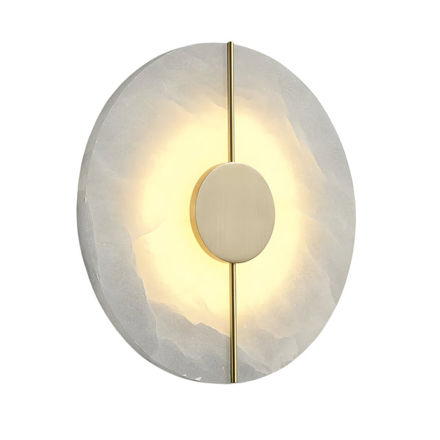 The Elaris LED Round Alabaster Wall Light in Soft White & Brass features a circular alabaster base, central brass disc, and vertical brass rod, softly glowing from behind to cast a radiant halo on the stone surface.