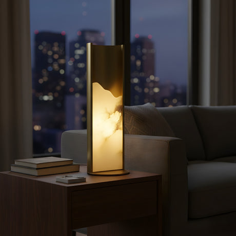 The Veloris LED Carved Alabaster Table Lamp in Brushed Brass & Soft White casts a warm glow on a wooden side table with books, set next to a sofa and large windows framing a city skyline at dusk.