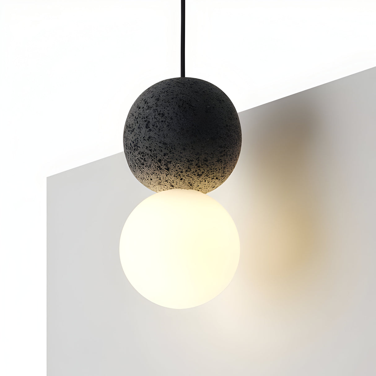 The Origo Concrete Pendant Light - Black & White is a minimalist fixture with a spherical design that features one half resembling dark, textured volcanic rock and the other crafted from smooth, opal glass. Suspended against a plain white background, it casts a gentle light.