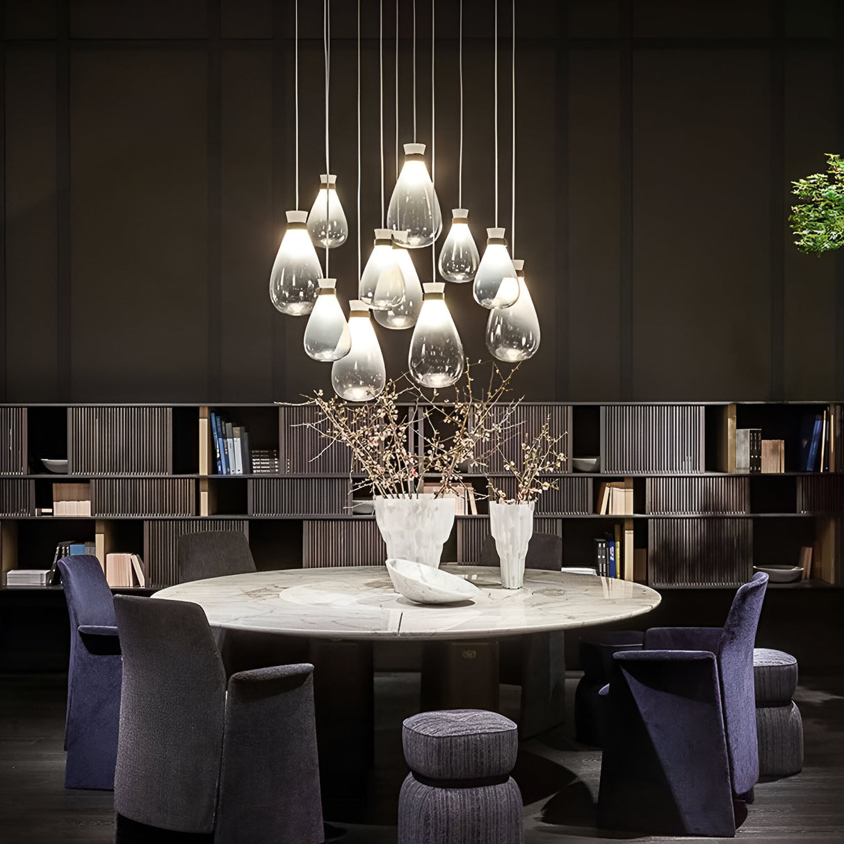 A contemporary dining room showcases a round marble table, encircled by blue upholstered chairs and matching ottomans. Above, a cluster of Soffi Small Glass Pendant Lights in clear glass hang from the ceiling, reflecting a handcrafted design aesthetic. Two white vases with branches adorn the table, while a bookshelf lines the wall.