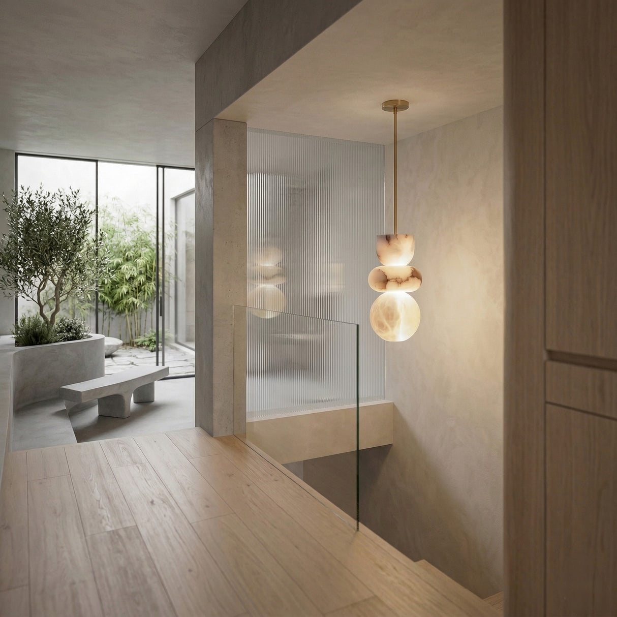 Modern minimalist interior with light wood floors, glass stair railing, and large windows opening to a courtyard with greenery and concrete benches. The space is illuminated by the Monne LED Large Stacked Alabaster Single Pendant Light in Soft White & Brushed Brass.