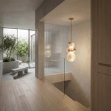 Modern minimalist interior with light wood floors, glass stair railing, and large windows opening to a courtyard with greenery and concrete benches. The space is illuminated by the Monne LED Large Stacked Alabaster Single Pendant Light in Soft White & Brushed Brass.