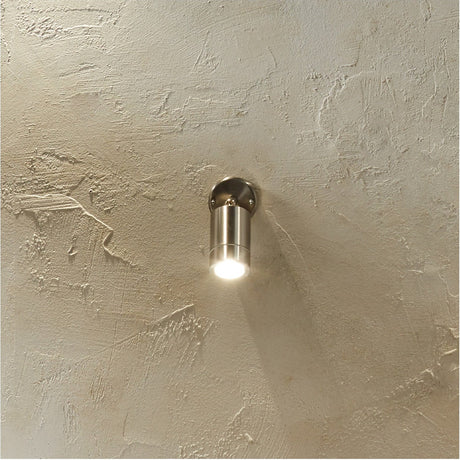 Lantana brushed steel adjustable directional spot wall light, 9cm cylindrical fitting mounted on a textured plaster wall, casting a downward beam of light.