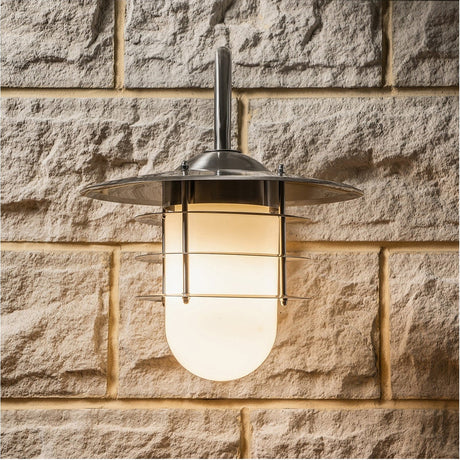 Centauri Outdoor Wall Light in brushed steel with an opaque glass shade and caged cover, mounted on a stone wall, illuminated.