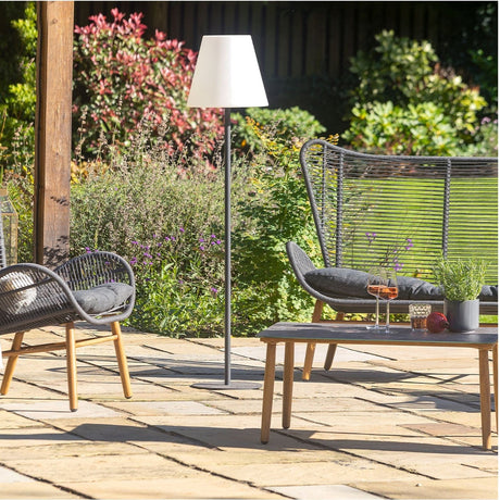 Grey Outdoor Floor Lamp, 153cm, with a slim aluminium pole and tapered white shade, shown on a garden patio beside woven outdoor chairs.