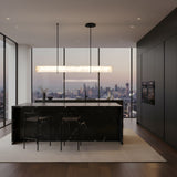 A modern kitchen with dark cabinets, a marble island, and city views at dusk is illuminated by the Zarune LED Linear Drop Alabaster Suspension Light in Matte Black & Soft White, adding an elegant touch above two bar stools.
