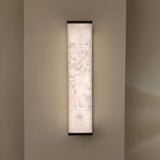 Eryndor LED Alabaster Wall Light in soft white stone with matt black end caps, illuminated on a neutral wall showing its tall rectangular panel and recessed light channel.