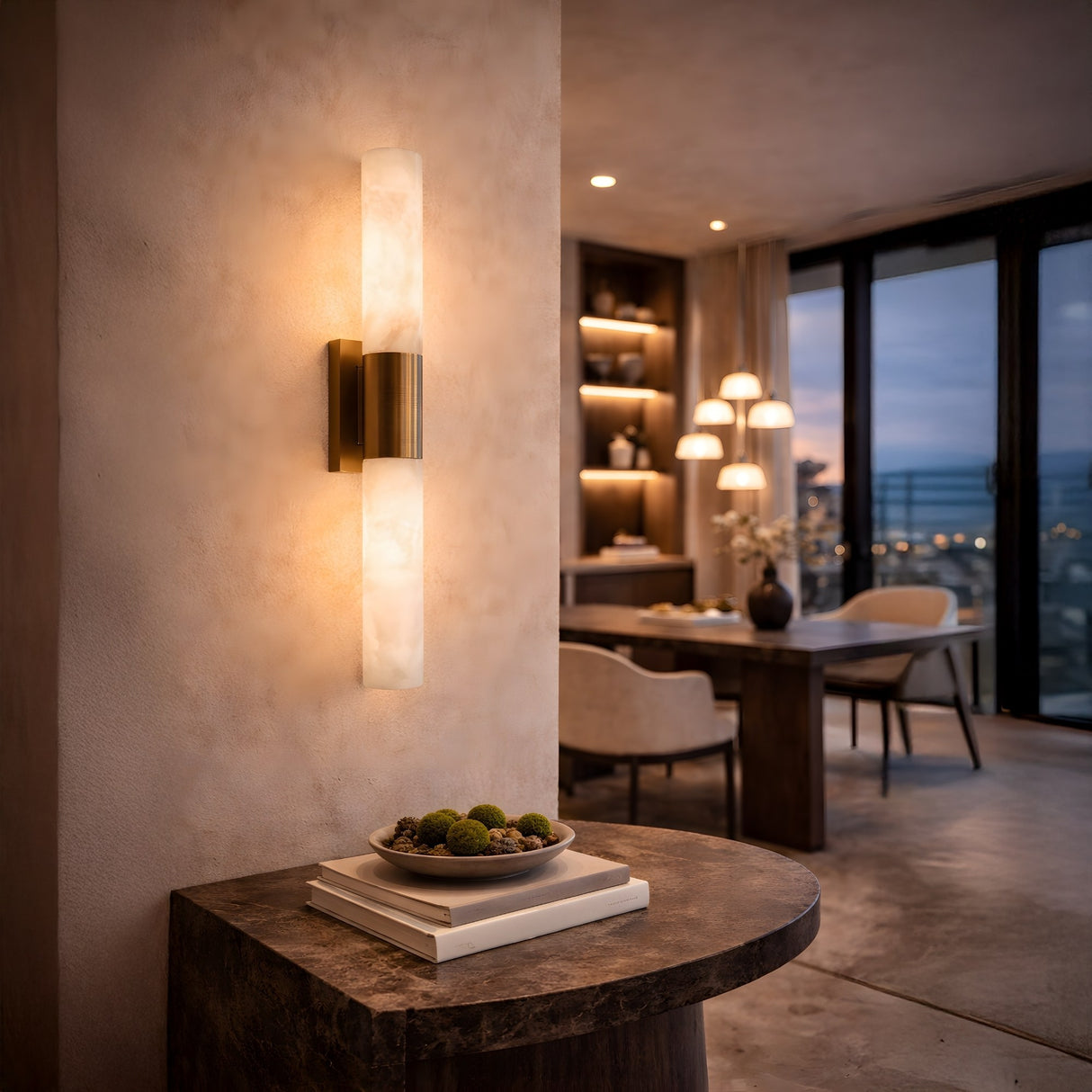 The Eryon LED Small Alabaster Wall Light in brushed brass and soft white glows warmly above a console with books and a bowl, while contemporary dining furniture frames a city view at dusk in the background.